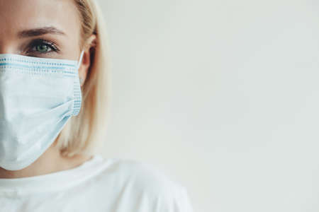 Close photo of a stunning caucasian woman with blonde hair wearing a medical mask and white t-shirt on a studio wall with free spaceの写真素材
