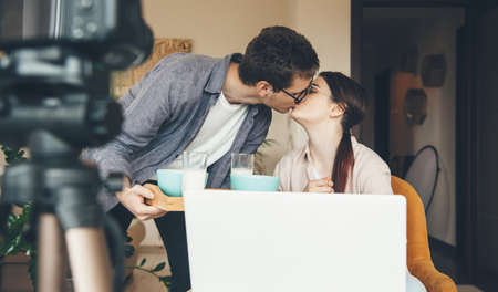 Charming caucasian woman with eyeglasses is having an online course while her man brings her the breakfastの写真素材