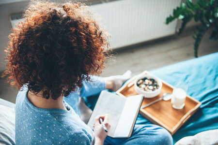 Upper view photo of a caucasian curly haired woman with healthy habits reading a book in bed and eating cereals with milkの写真素材
