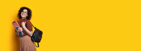 Charming lady with curly hair and eyeglasses holding a bag and folders on a yellow studio wall with...