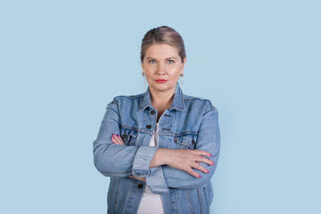 Serious senior caucasian woman with blonde hair wearing jeans coat posing with crossed hands on a...