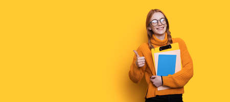 Lovely red haired student with freckles gesturing the like sign and holding folders on a yellow wall...