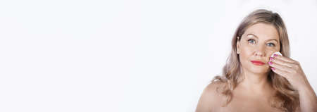 Adult caucasian blonde woman cleaning her face with a cotton disk posing with naked shoulders on a...