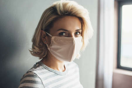 Caucasian woman with blonde hair and mask looking at camera and smile posing inside near the windowの写真素材