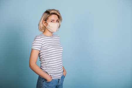 Charming blonde lady wearing a white protective mask posing on a blue studio wall with free spaceの写真素材
