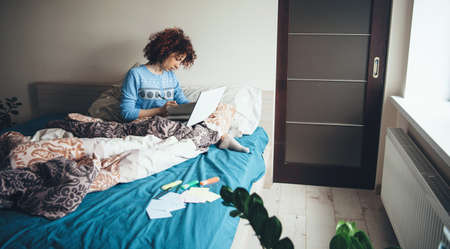 Charming lady with curly hair wearing a blue pajama is using a laptop in bed while doing homework