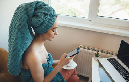 Caucasian woman with a towel on head is chatting on mobile and laptop at home after the bathの写真素材