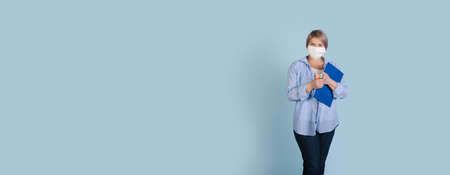 Senior caucasian businesswoman with medical mask on face holding a folder and markers is posing on a blue studio wall with free spaceの写真素材