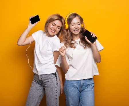 Two blonde women listening to music with headphones and mobile on yellow studio wallの写真素材