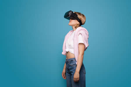 Young blonde woman using virtual reality headset and posing on a blue studio wall