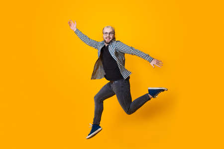 Caucasian man with blonde hair and eyeglasses jumping on a yellow studio wall while listening to...
