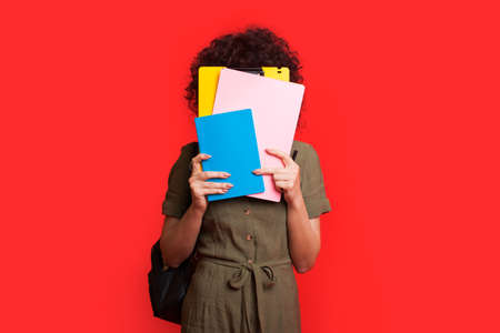 Caucasian student with curly hair covering her face with books and posing on a red studio wall holding a bagの写真素材
