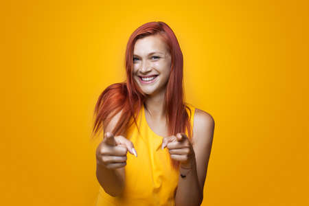 Monochrome photo of a ginger woman pointing at camera and smile wearing a yellow dress on studio...