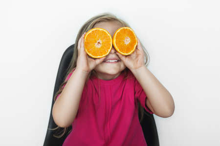 Blonde small girl with healthy habits is covering her eyes with a sliced orange and smile on a white studio wallの写真素材