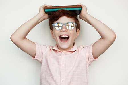 Young caucasian boy with ginger hair holding some book s on his head and smile at camera wearing...
