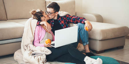 Lovely caucasian couple sitting on the floor and kissing while using a laptop and holding an orange covered with a quiltの写真素材