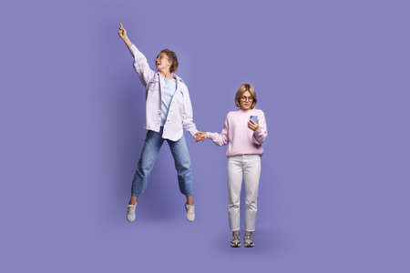 Two caucasian women posing on a violet studio wall pointing up and jumping while chatting on mobile...