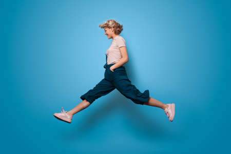 Charming blonde lady jumping on a blue studio wall and smile advertising somethingの写真素材