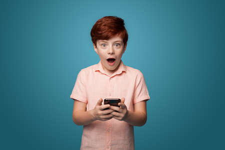 Shocked caucasina ginger boy holding a phone and looking at camera on a blue studio wallの写真素材