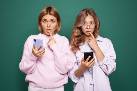 Two disappointed blonde women chatting on a green studio wall is touching her lips with finger and look sad at cameraの写真素材