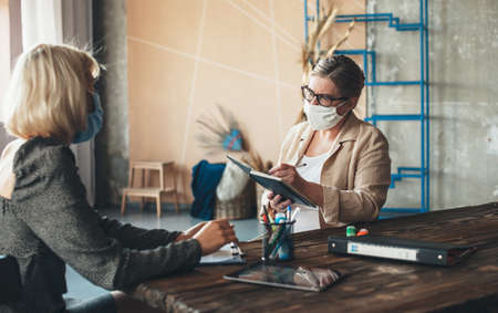 Serious caucasian senior woman with medical mask on face and glasses is discussing with a client at home while write something in the bookの写真素材