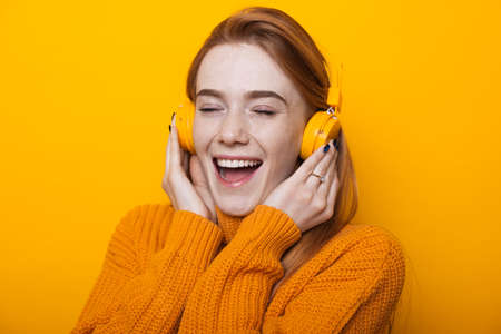 Close up photo of a ginger woman with freckles listening to music using headphones on a yellow studio wallの写真素材