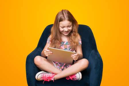 Cute small girl playing with a tablet sitting in the chair on a yellow studio wall