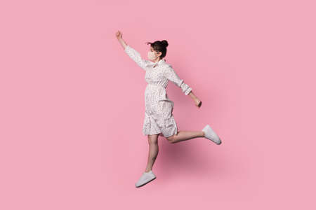 Cute brunette lady jumping on a pink studio wall gesturing like a superwoman and wear a medical mask...
