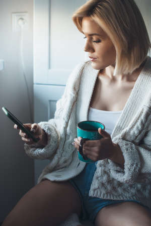 Close up photo of a blonde charming lady in knitted sweater sitting in the kitchen and drinking a...