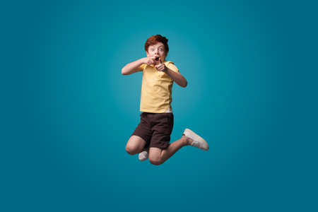 Ginger caucasian boy jumping on a blue studio wall gesturing and pointing at camera with forefinger and opened mouthの写真素材