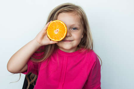 Small blonde girl covering her eye with a sliced orange posing in a red shirt on a white studio wall smiling at cameraの写真素材