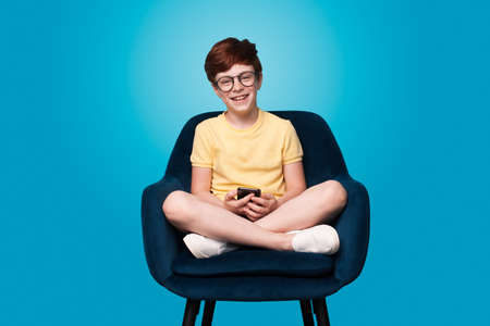 Ginger boy sitting in armchair and wear eyeglasses is chatting on phone posing on a blue studio wall