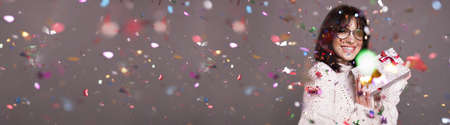 Caucasian brunette woman with eyeglasses is posing with a christmas present at camera on a gray studio wall full of confettiの写真素材