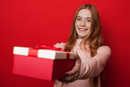 Caucasian woman with freckles and ginger hair is giving a box with gift at camera and smile on red studio wallの写真素材