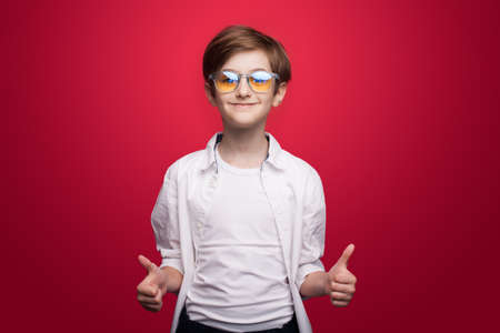 Pleased caucasian boy with glasses and white shirt gesturing like sign and smile at camera on a red studio wallの写真素材