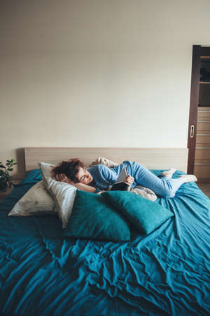 Curly haired lady in blue pajama is reading a book in bed enjoying weekend