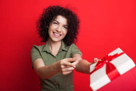 Charming curly haired woman smiling and giving a present at camera gesturing with finger that is is for you while posing on a red studio wallの写真素材