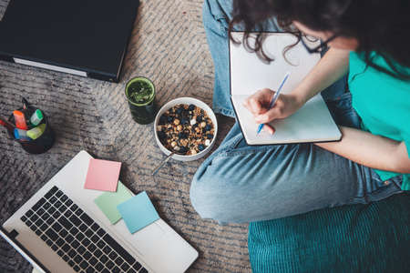 Upper view photo of a caucasian woman eating cereals while writing something in a book and using a computer on the floorの写真素材