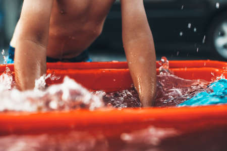 Caucasian undressed boy playing with hands is water at home with a toy and a basin in a summer dayの写真素材