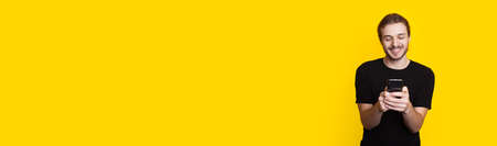 Long haired man with beard is chatting on mobile while smile in a black t-shirt on a yellow studio...