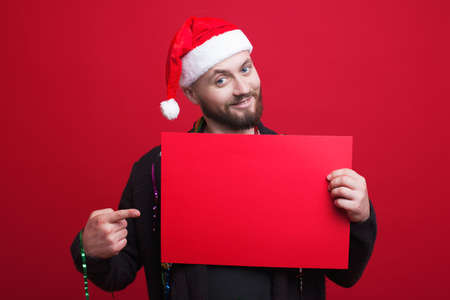 Young man with beard and new year hat is pointing to red blank space on a banner holding on a studio...