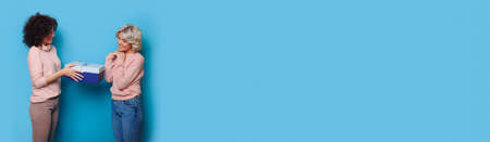 Blonde woman receiving a present from her brunette sister gesturing pleasure on a blue wall with...