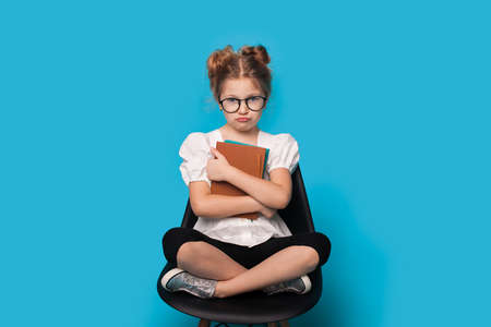 Funny caucasian school girl gesturing disappointment on a blue studio wall while wear glasses and sitting in armchair holding booksの写真素材