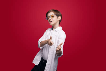 Cute caucasian boy with glasses is pointing and blinking at camera posing on a red studio wallの写真素材