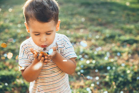Small boy blowing confetti from hand while walking outside on the grass in a sunny day