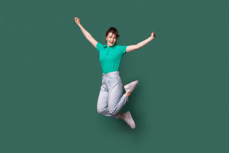 Brunette caucasian woman in jeans and t-shirt jumping on a green studio wall and smile