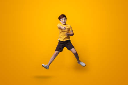 Jumping caucasian boy with red hair is wearing glasses and point at camera wearing a yellow shirt on a studio wallの写真素材