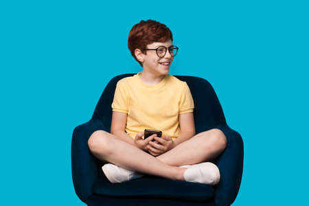 Ginger caucasian boy smiling and wear glasses on a blue studio wall while holding and using a mobileの写真素材
