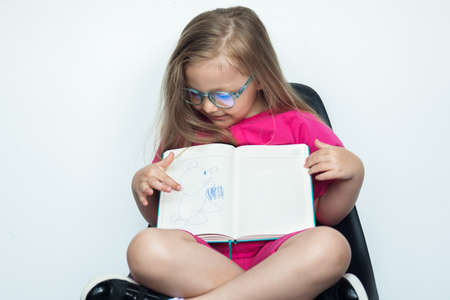 Small caucasian girl with blonde hair and glasses sitting in armchair on a white studio wall is...