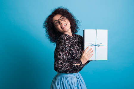 Curly haired woman with glasses wearing a dress is presenting at camera a box with gift posing on a blue studio wallの写真素材